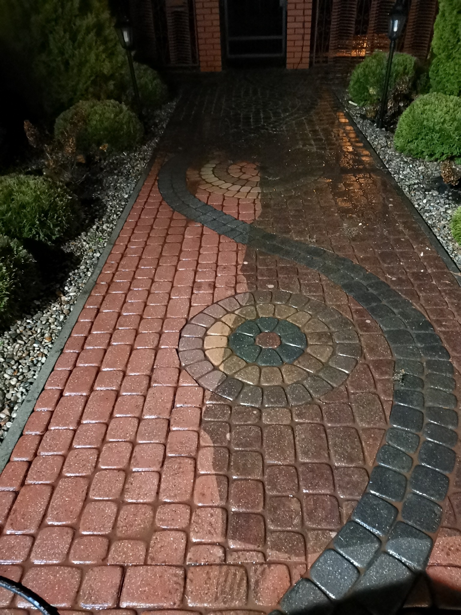 Mokry chodnik z czerwonej kostki brukowej z dekoracyjnym wzorem w kształcie spirali i koła, oświetlony lampami ogrodowymi, otoczony zielenią i kamieniami.