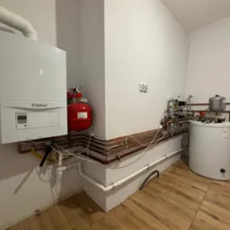 Biały piec gazowy Vaillant zamontowany na ścianie, połączony z czerwoną butlą i siecią miedzianych rur, obok biały zasobnik ciepłej wody i liczne zawory, całość na tle białych ścian i brązowej...