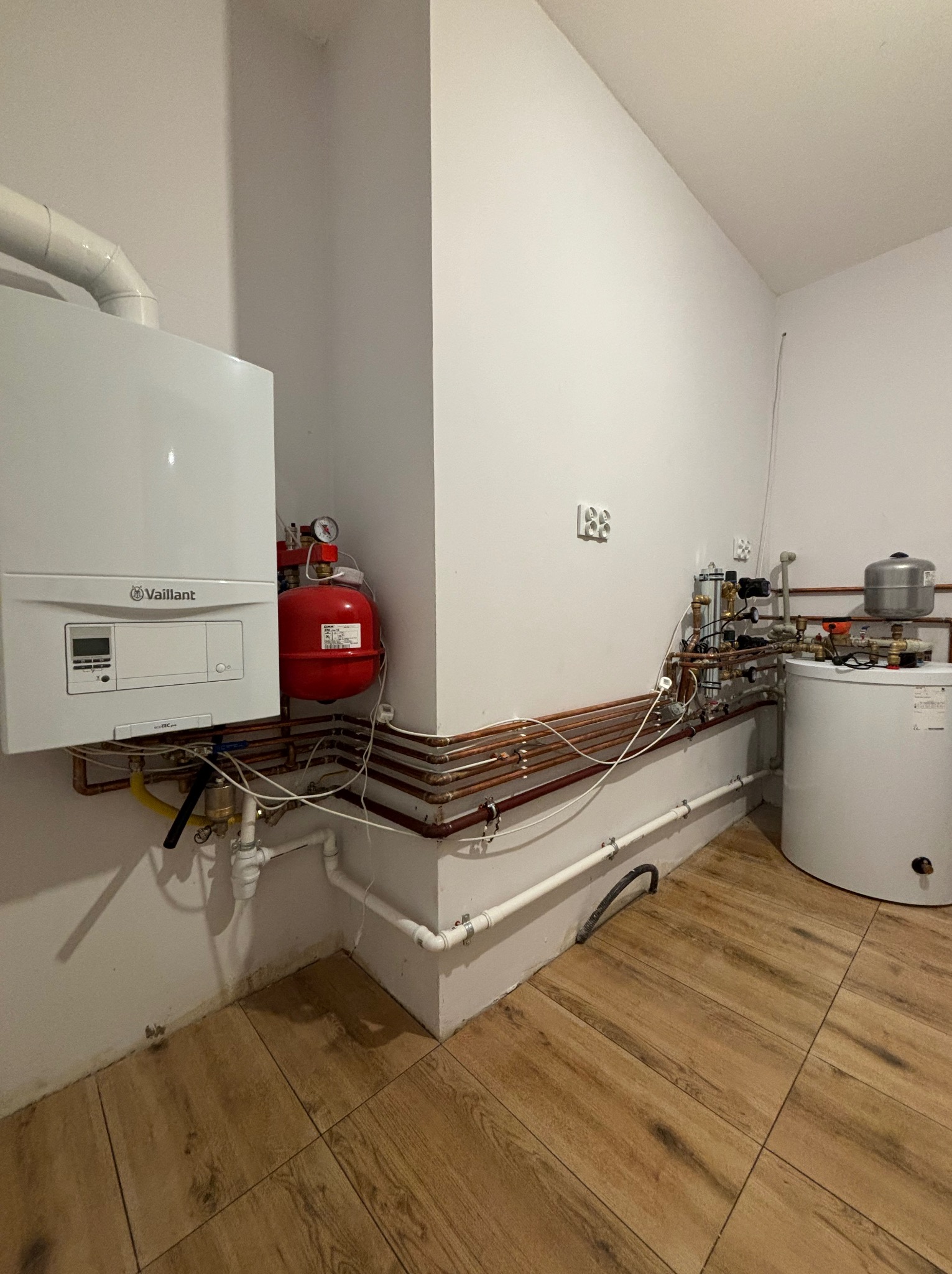 Biały piec gazowy Vaillant zamontowany na ścianie, połączony z czerwoną butlą i siecią miedzianych rur, obok biały zasobnik ciepłej wody i liczne zawory, całość na tle białych ścian i brązowej...