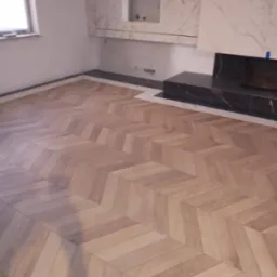 Nowo ułożony parkiet w jodełkę klasyczną, jasne drewno, widoczne fragmenty ściany z marmurowym wykończeniem kominka, w tle okno.