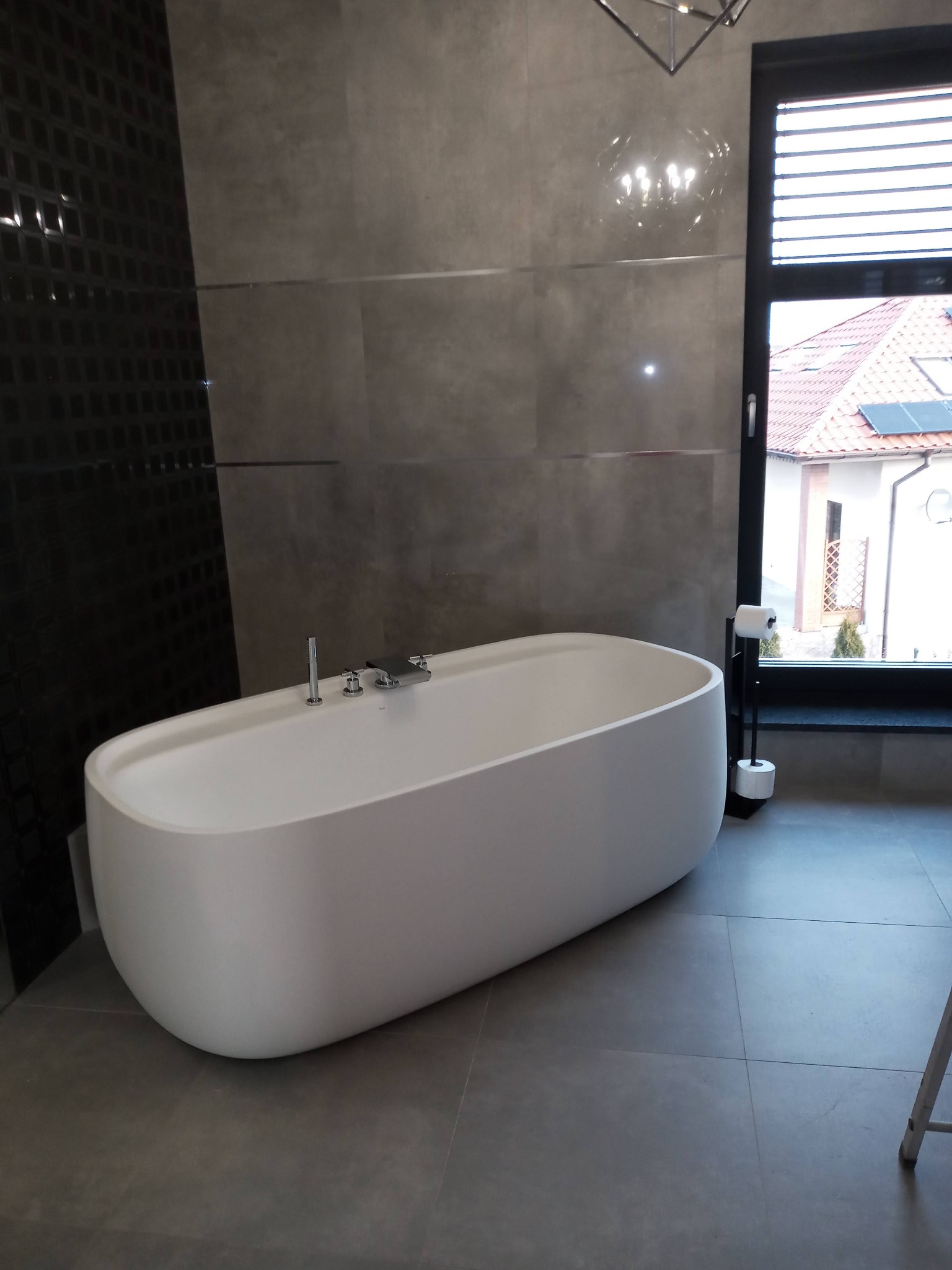 Nowoczesna łazienka z wolnostojącą białą wanną na tle szarych płytek i czarnej ściany z dekoracyjnymi elementami, widok na dom przez okno.