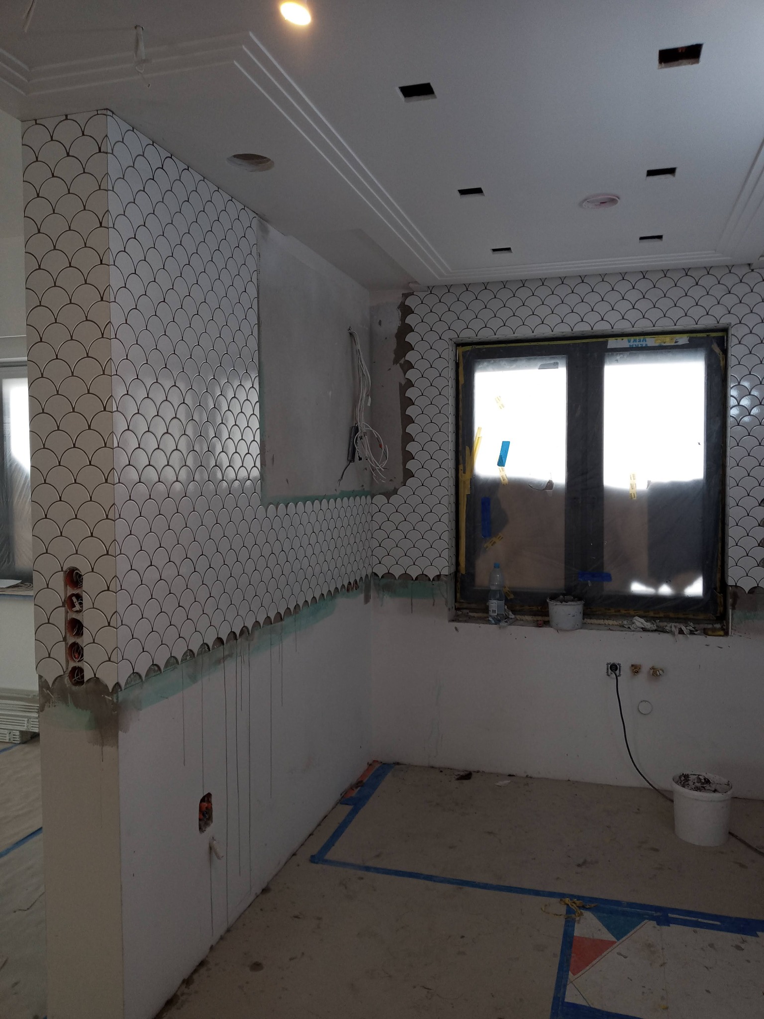 Ściana w trakcie wykańczania białymi płytkami w kształcie rybiej łuski, widoczne niedokończone fugi, okno zabezpieczone folią, przygotowane gniazdka elektryczne.