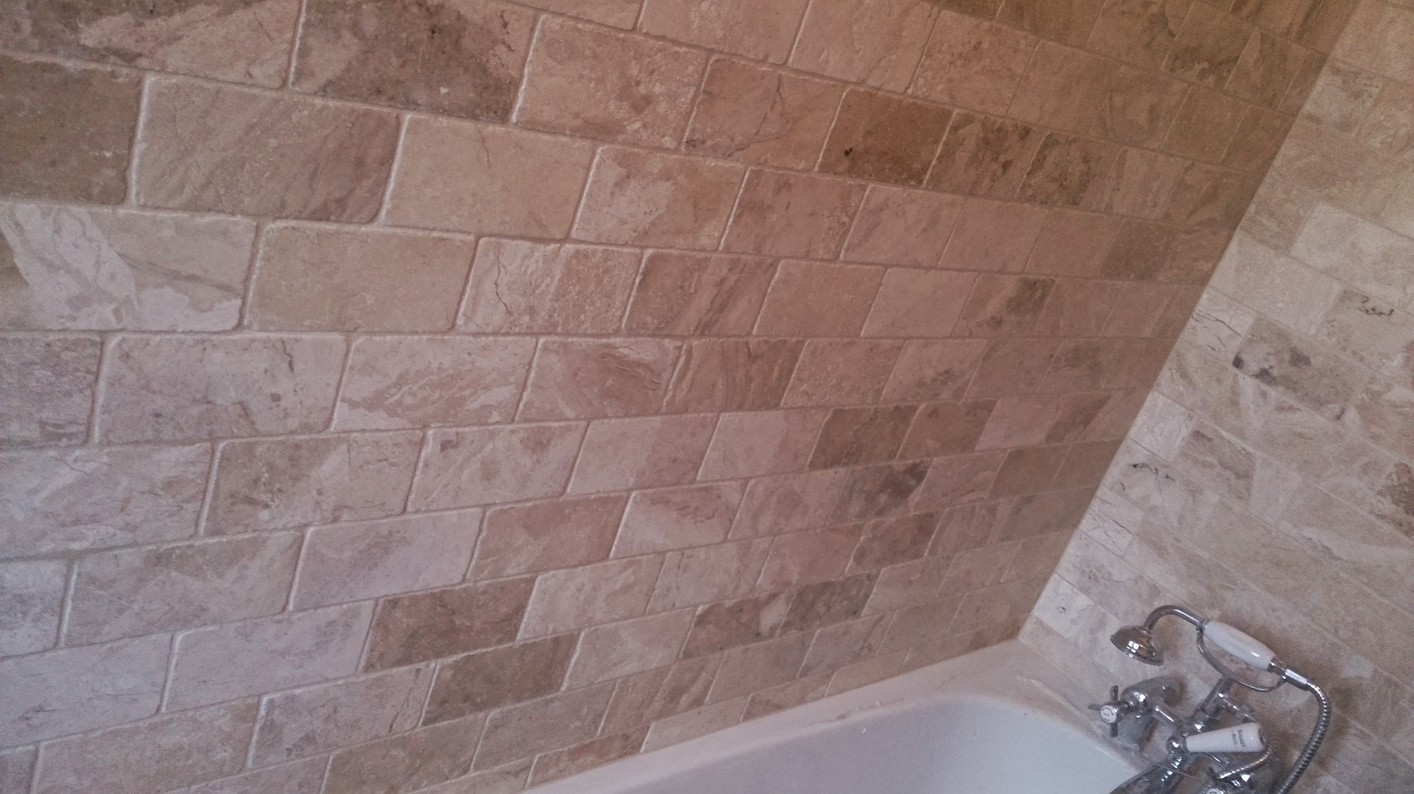 Ściana łazienki wyłożona beżowymi, marmurkowymi płytkami ceramicznymi ułożonymi w cegiełkę, widoczna bateria wannowa z chromowanymi kurkami.