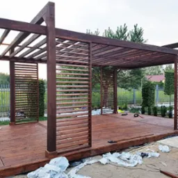 Drewniana konstrukcja zadaszenia z lamelami na podeście, otoczona zielenią, w trakcie montażu, widoczne narzędzia i materiały budowlane.