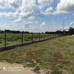 Długi, szary fundament betonowy z czarnym ogrodzeniem panelowym na tle zielonego pola i błękitnego nieba z chmurami.