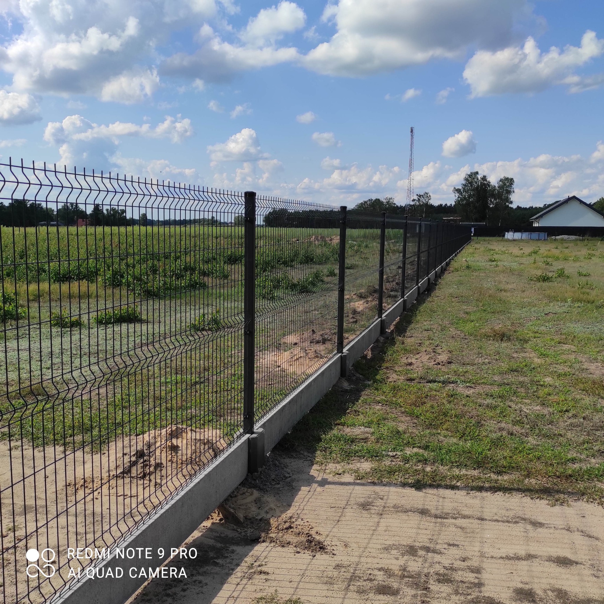 Długi, ciemny, metalowy płot panelowy na betonowej podmurówce, rozciągający się wzdłuż granicy posesji na tle zielonego pola i błękitnego nieba z chmurami.