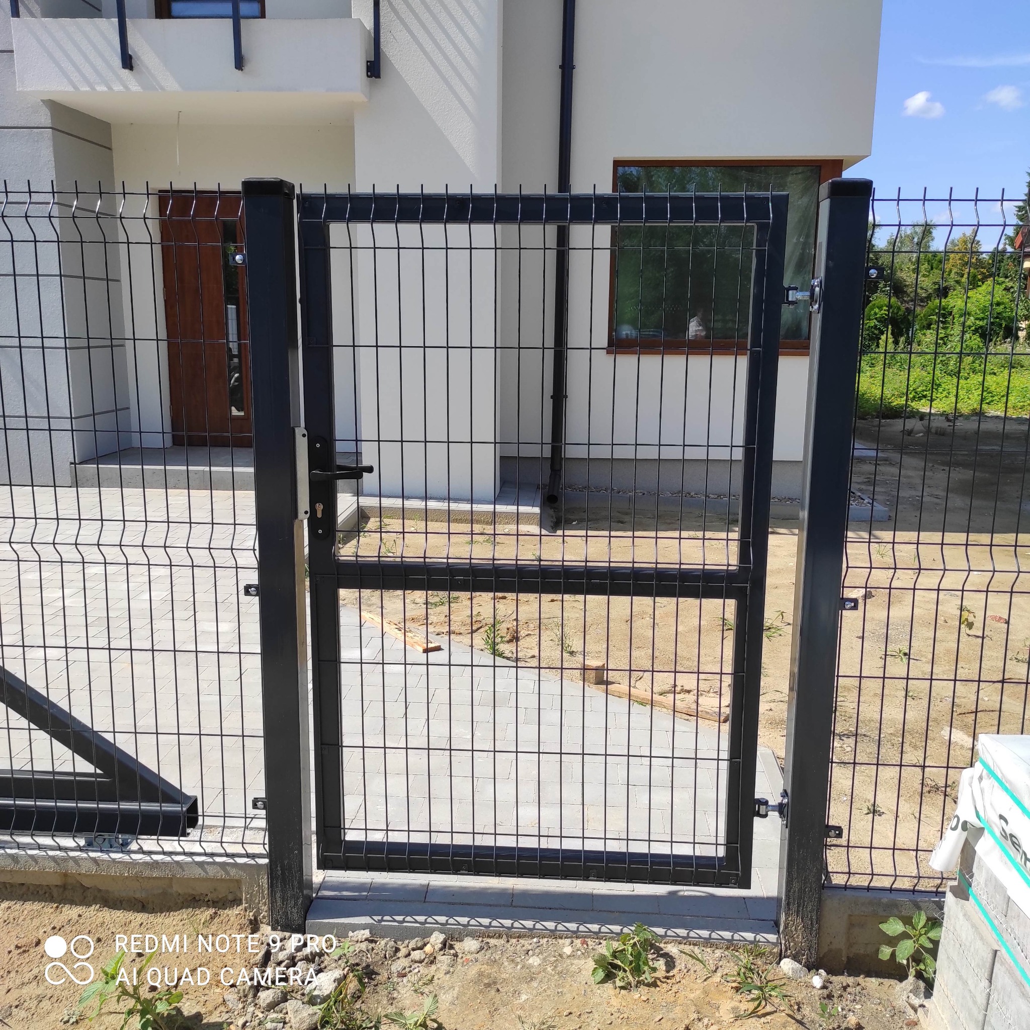 Nowoczesna brama panelowa w kolorze czarnym, część ogrodzenia posesji w Głownie, z widocznym fragmentem domu w tle.