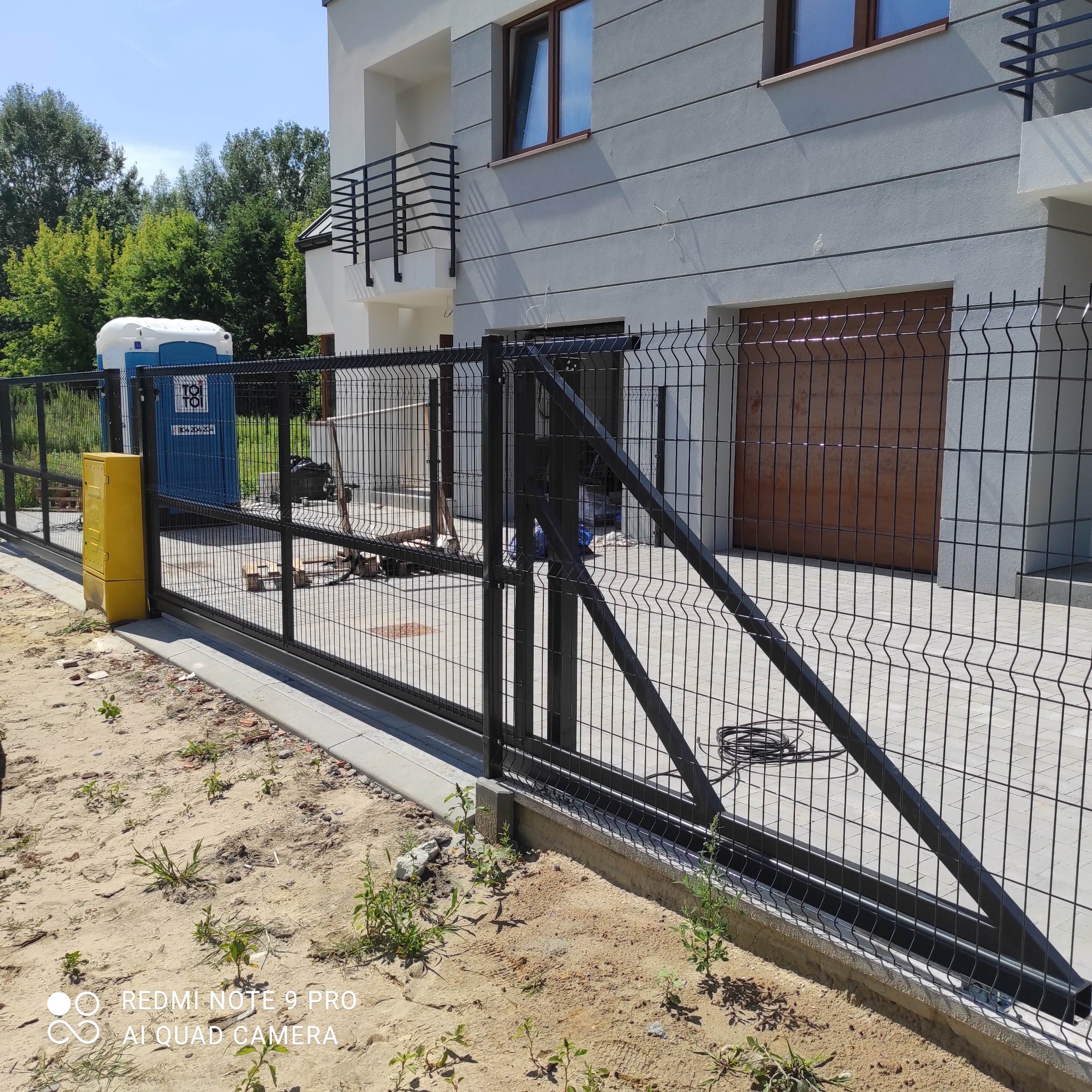 Nowoczesne ogrodzenie panelowe w kolorze czarnym, z przesuwną bramą, na tle jasnego domu z brązową bramą garażową; widoczna przenośna toaleta TOI TOI.