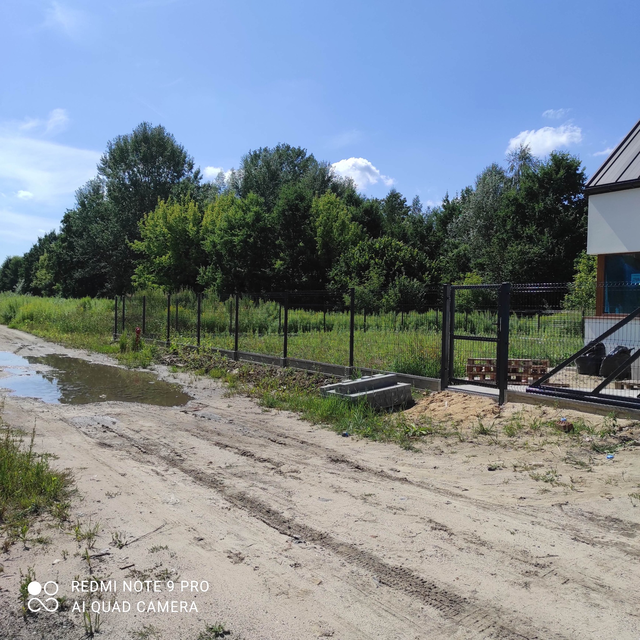 Nowo postawione ogrodzenie panelowe z bramą wjazdową, widoczne betonowe podmurówki i słupki, w tle drzewa i fragment domu w słoneczny dzień, na pierwszym planie błotnista droga.
