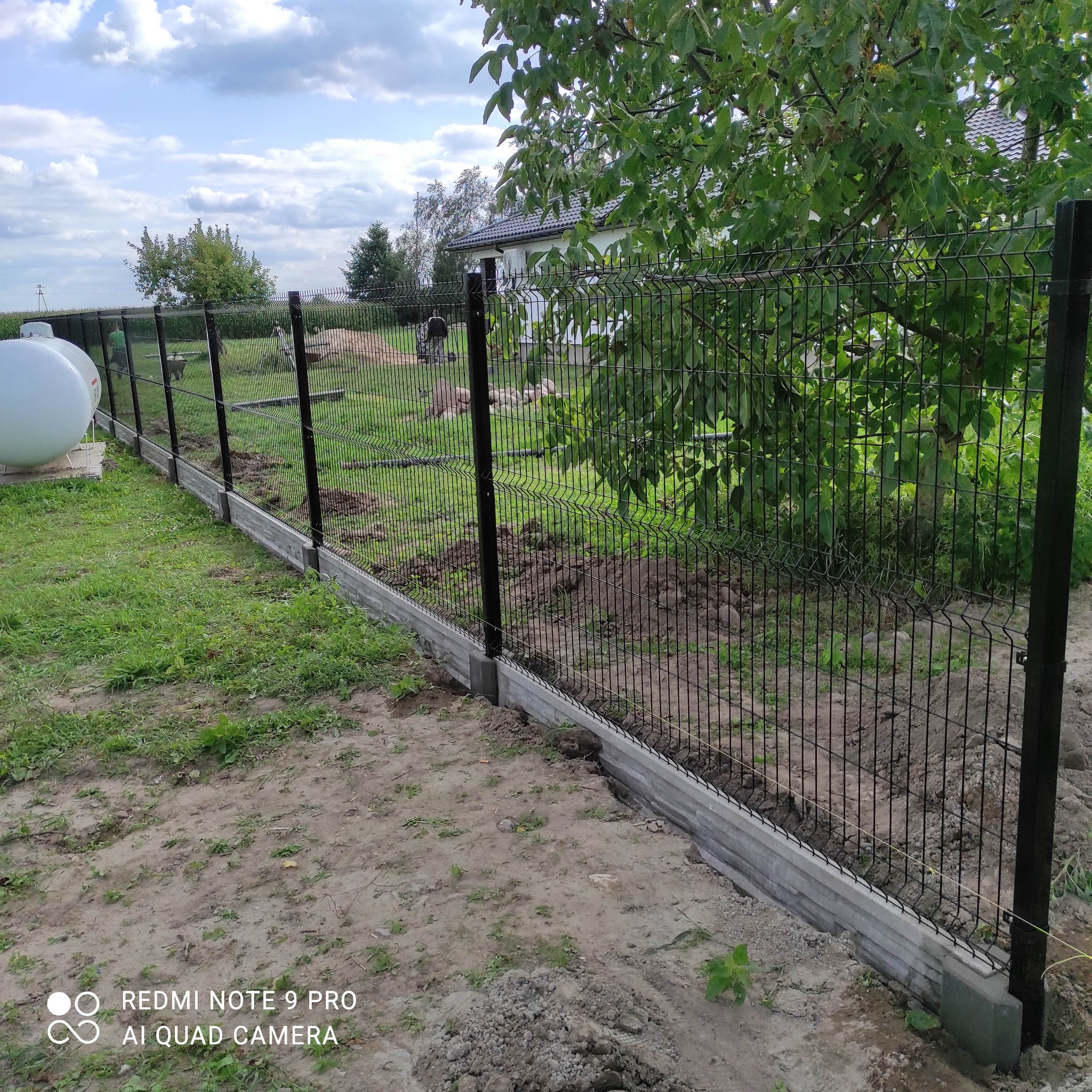 Nowo zamontowane ogrodzenie panelowe z podmurówką betonową, widoczne zbiorniki na gaz w tle, teren jeszcze niezagospodarowany.