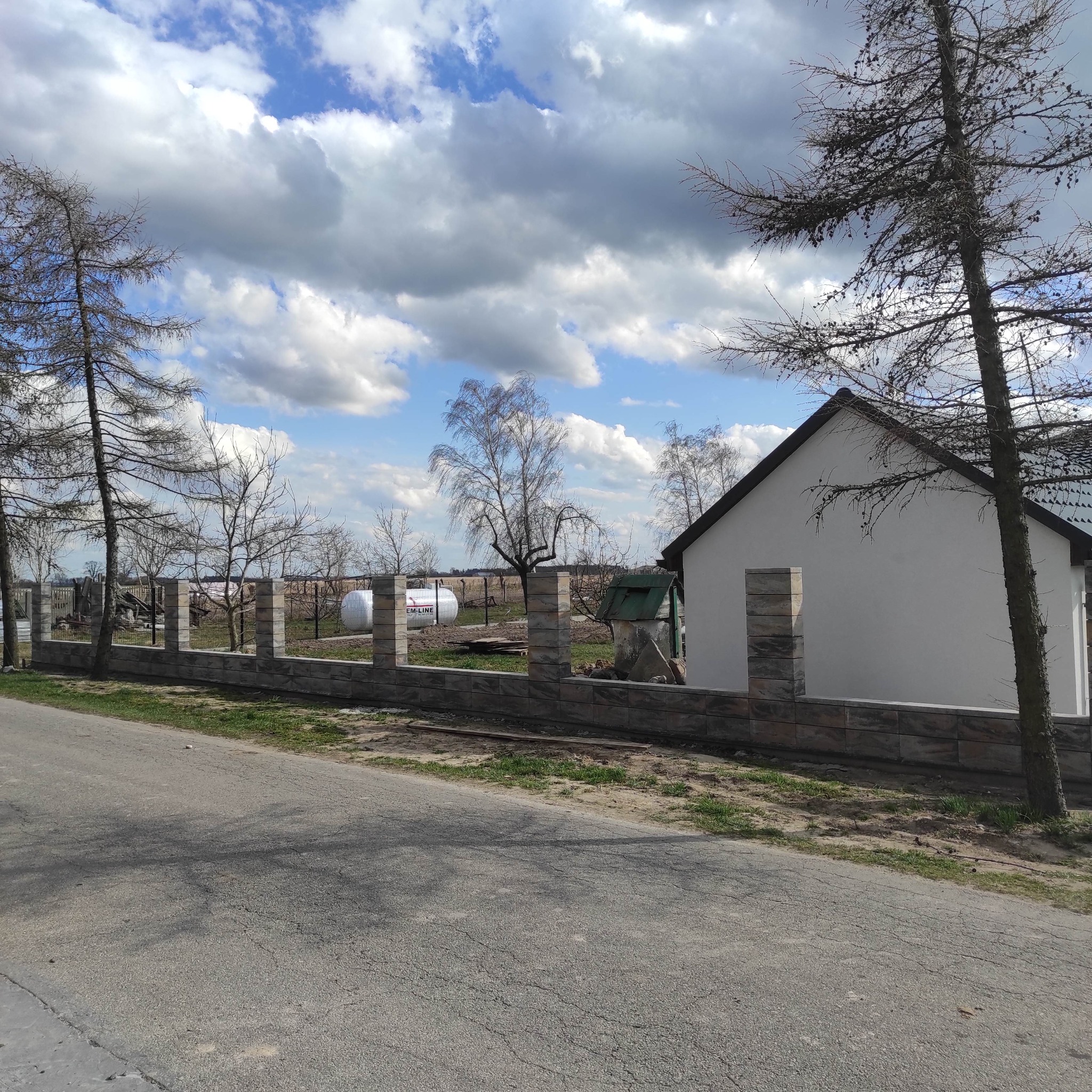 Niski murek z bloków imitujących kamień, tworzący ogrodzenie posesji z widocznym zbiornikiem gazu w tle, na tle pochmurnego nieba.