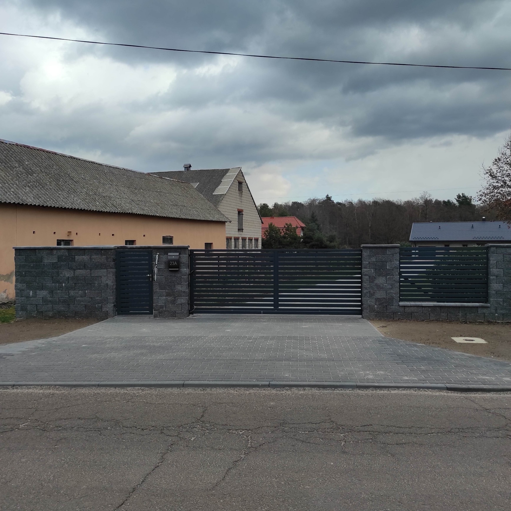 Nowoczesna, pozioma brama przesuwna z szarego metalu, osadzona między słupkami z szarej kostki, prowadząca na wybrukowany podjazd, na tle pochmurnego nieba i zabudowań.