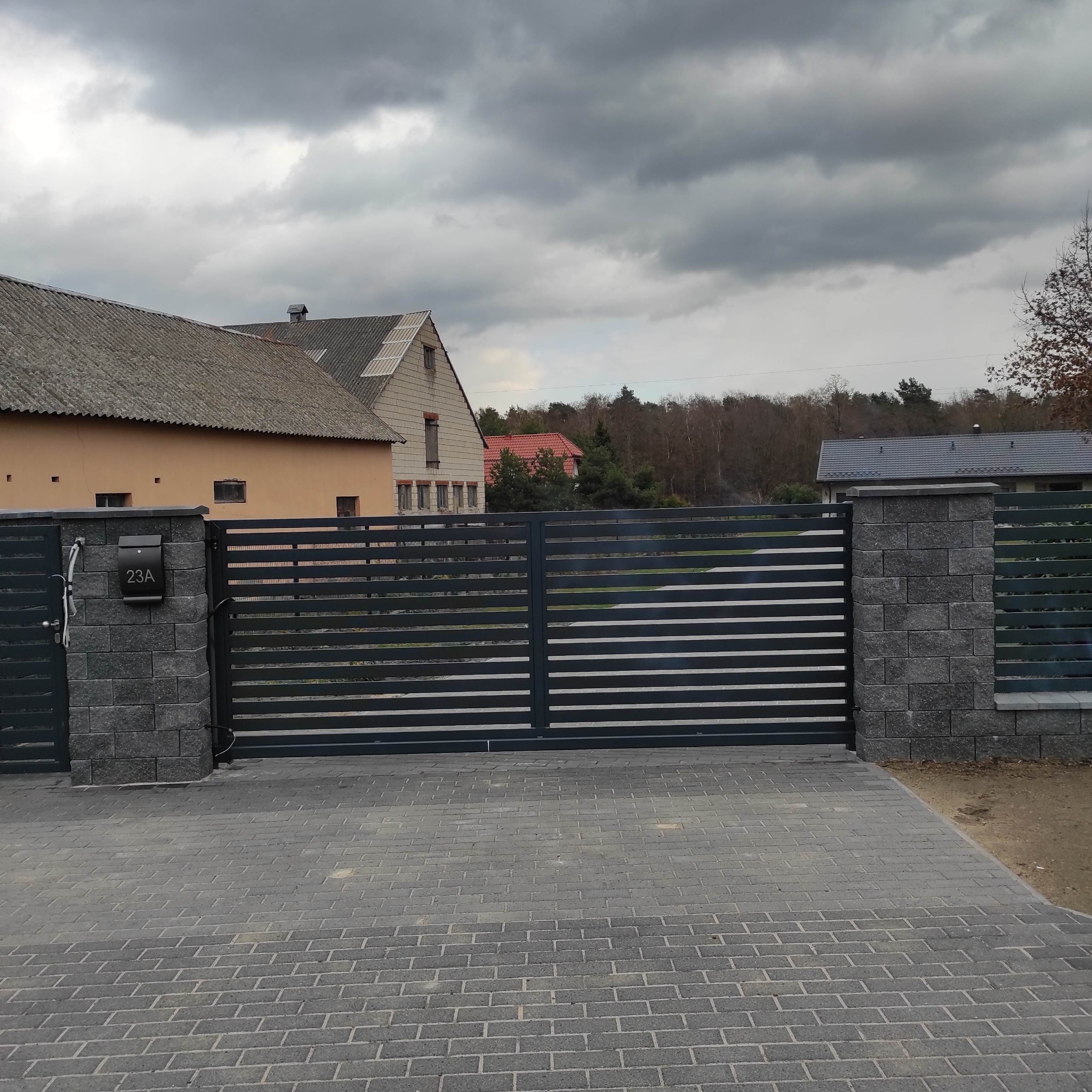 Nowoczesna, pozioma brama wjazdowa z metalowymi przęsłami, osadzona na słupkach z szarej kostki, z numerem domu 23A na skrzynce pocztowej, na tle budynków mieszkalnych i pochmurnego nieba.