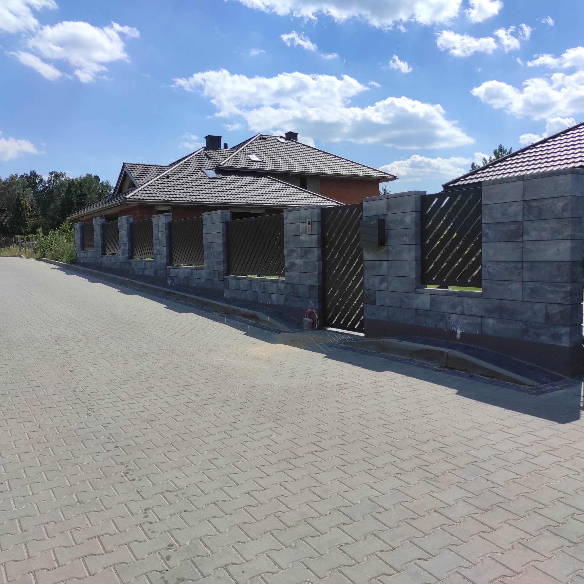 Nowoczesne ogrodzenie z szarego betonu architektonicznego z metalowymi przęsłami o ukośnym wzorze, widok na dom z ciemną dachówką i kostkę brukową w słoneczny dzień.