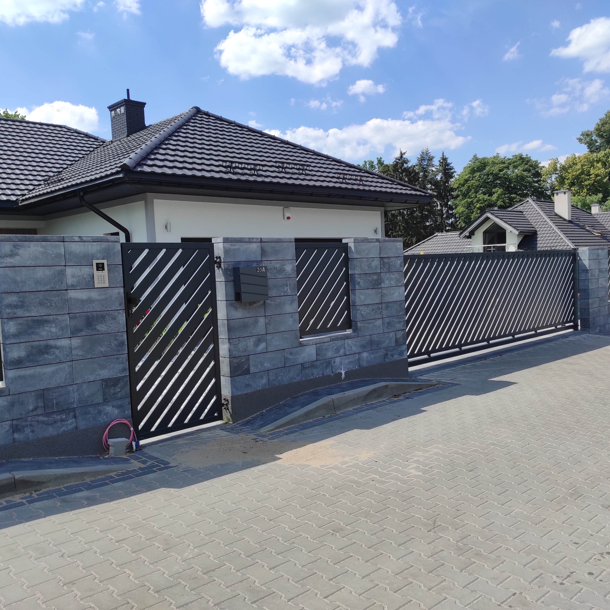 Nowoczesne ogrodzenie posesji z betonowych bloczków i metalową bramą przesuwną z ukośnymi szczeblami, widok z zewnątrz na tle domu jednorodzinnego i błękitnego nieba.