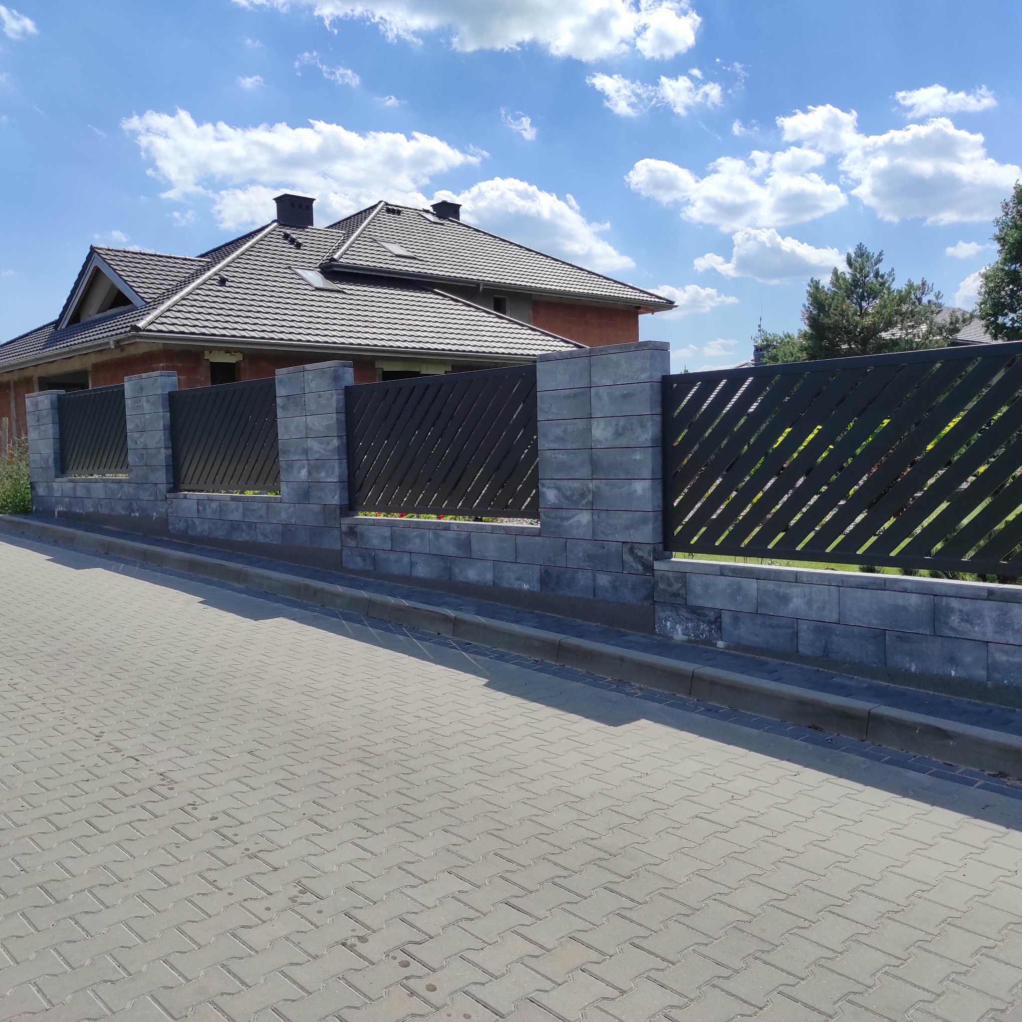 Nowoczesne ogrodzenie z szarej kostki betonowej i metalowych paneli z ukośnymi szczeblami, widok na dom z ciemnobrązową dachówką i błękitne niebo z białymi chmurami.