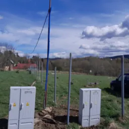 Dwa szare, bliźniacze słupy elektryczne z żółtymi znakami ostrzegawczymi na zielonym trawiastym polu, z metalowymi słupkami w tle i niebieskim samochodem w oddali pod błękitnym niebem z chmurami.