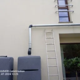 Szara rynna spustowa z odpływem do zbiornika na deszczówkę, drabina oparta o ścianę budynku, okno z metalowymi wspornikami.