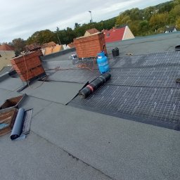 ADAM PIETRZAK USŁUGI OGÓLNOBUDOWLANE - Montaż papy termozgrzewalnej na dachu płaskim, widoczny pracownik w roboczych spodniach i obuwiu, butla gazowa, rolka papy oraz komin z cegły w tle zabudowań miejskich.