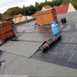 ADAM PIETRZAK USŁUGI OGÓLNOBUDOWLANE - Widok z góry na dach w trakcie pokrywania papą termozgrzewalną z widocznymi dwoma kominami z cegły, butlą gazową, narzędziami i rolką papy. Na dachu widoczne są również inne budynki.