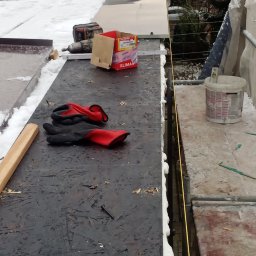 ADAM PIETRZAK USŁUGI OGÓLNOBUDOWLANE - Prace dekarskie w zimowych warunkach: narzędzia, wkrętarka, puszka z wkrętami Klimas, rękawice robocze i poziomica na dachu pokrytym śniegiem, widoczne pianowanie krawędzi.