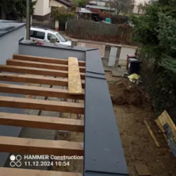 Konstrukcja dachu w trakcie budowy z widocznymi drewnianymi belkami i fragmentem obróbki blacharskiej w kolorze grafitowym, na tle zabudowań i zieleni.