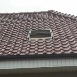 Czerwona dachówka ceramiczna z zamontowanym oknem dachowym, widoczne uszczelnienia i fragment elewacji budynku w tle.