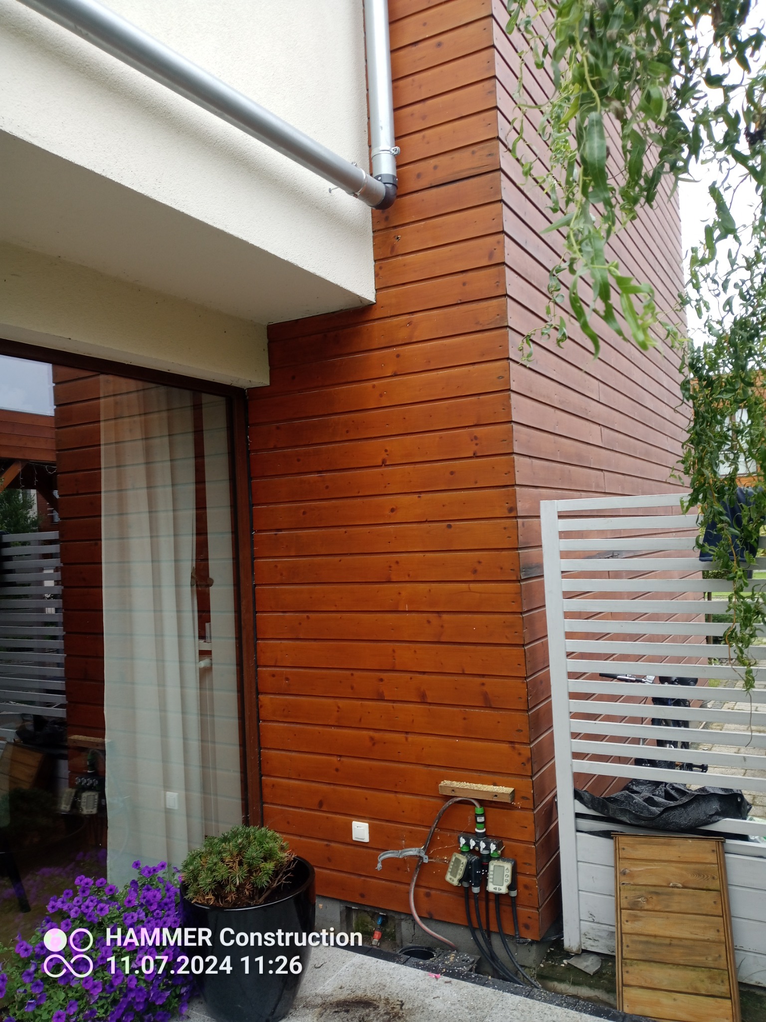 Fragment elewacji budynku wykończonej drewnianą boazerią, z widoczną rynną spustową, białym ogrodzeniem panelowym i instalacją nawadniającą przy ścianie.