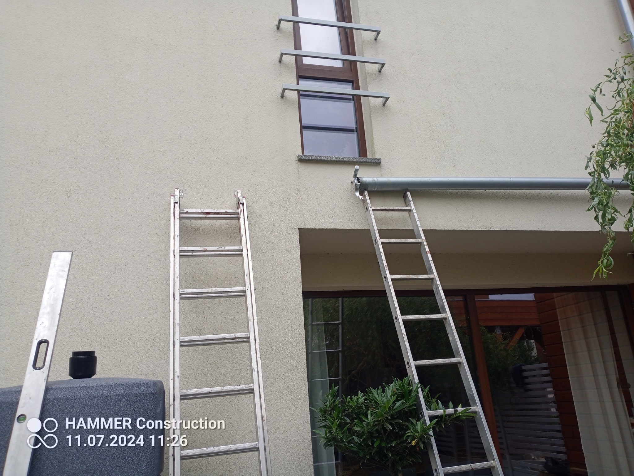 Przygotowania do prac na wysokości w Pniewach: dwie drabiny opierają się o elewację budynku, jedna przy oknie z zamontowanymi wspornikami, druga przy rynnie. Na pierwszym planie poziomica i czarny...