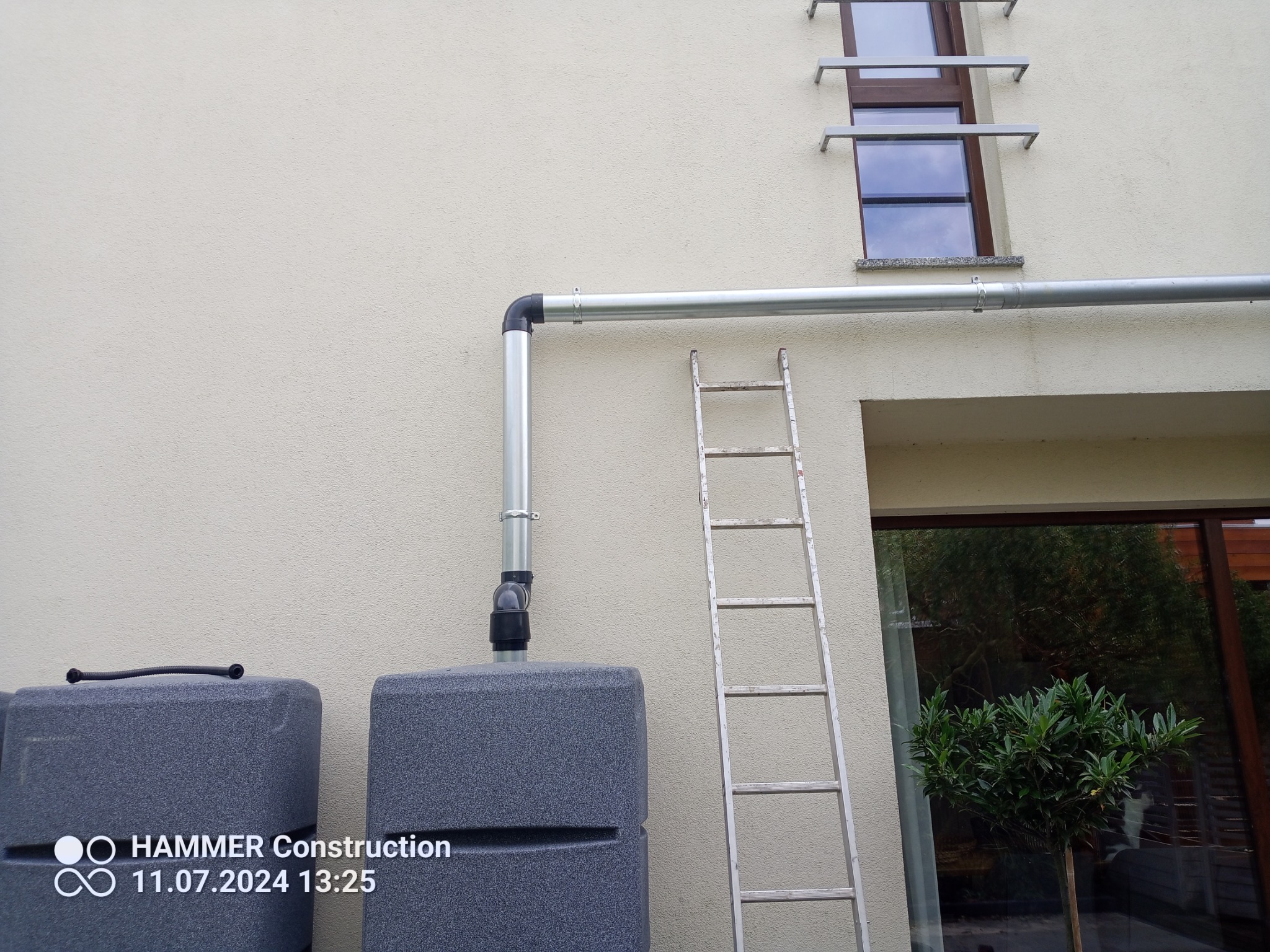 Szara rynna spustowa z odpływem do zbiornika na deszczówkę, drabina oparta o ścianę budynku, okno z metalowymi wspornikami.