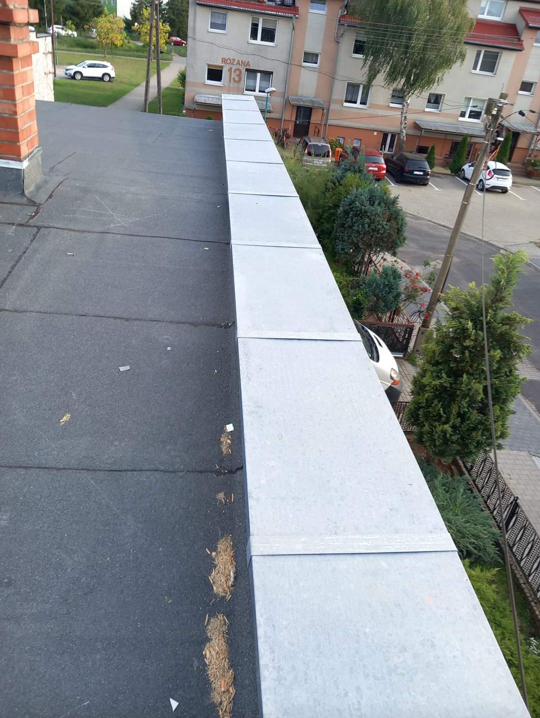 Widok z góry na płaski dach pokryty papą z nową, metalową obróbką blacharską krawędzi, w tle budynek mieszkalny z numerem 13 i zaparkowane samochody.