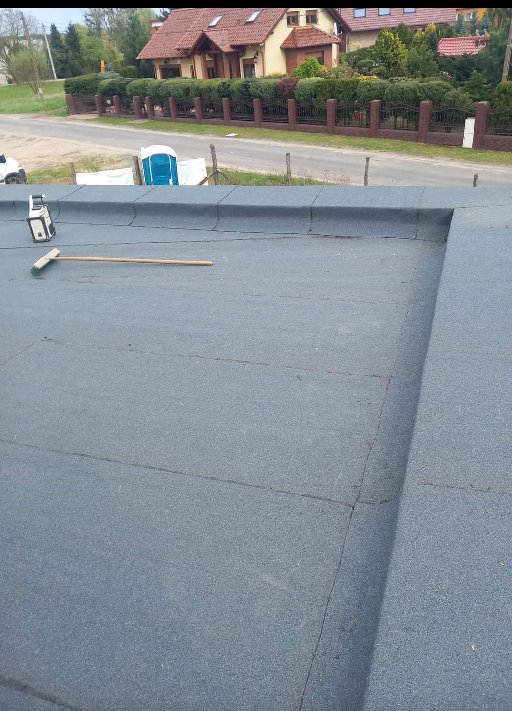 Widok z góry na płaski dach pokryty papą, z widocznym laserowym niwelatorem i miotłą, na tle domów jednorodzinnych i przenośnej toalety.