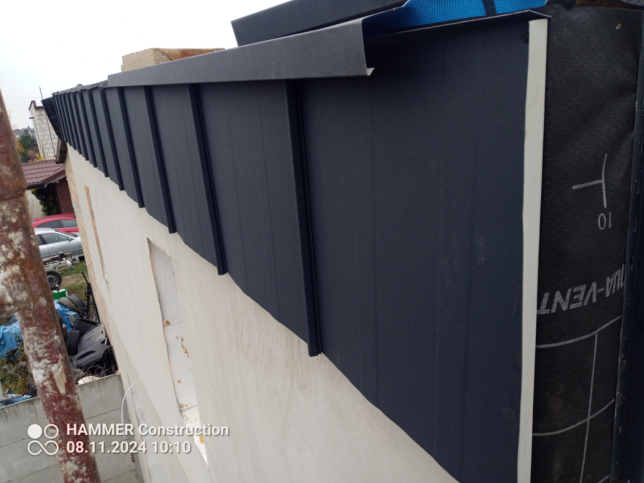 Fragment dachu z ciemnej blachy trapezowej, widoczna krawędź z obróbką blacharską, warstwa izolacyjna i membrana dachowa, ujęcie z dołu.