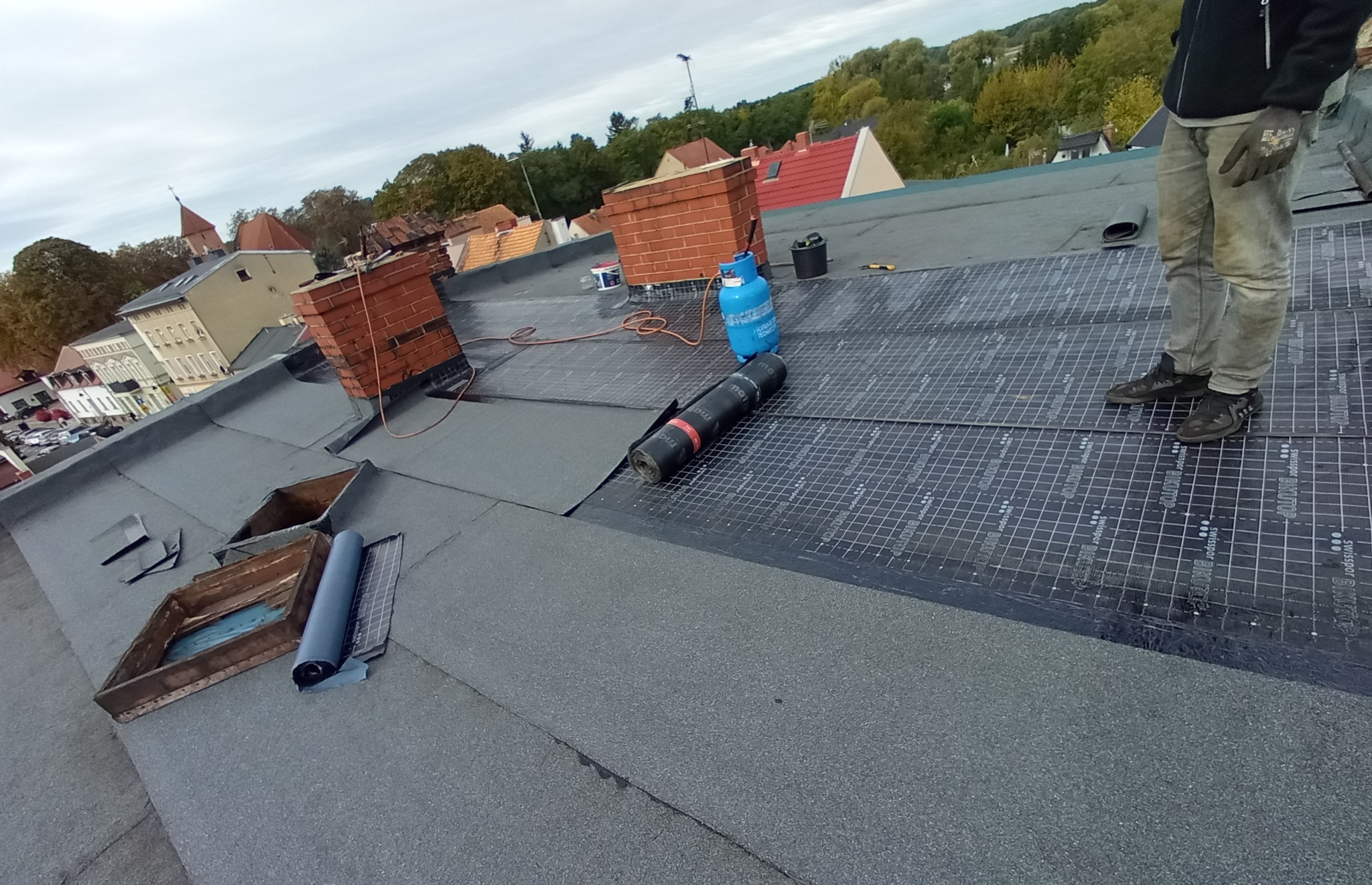 Montaż papy termozgrzewalnej na dachu płaskim, widoczny pracownik w roboczych spodniach i obuwiu, butla gazowa, rolka papy oraz komin z cegły w tle zabudowań miejskich.