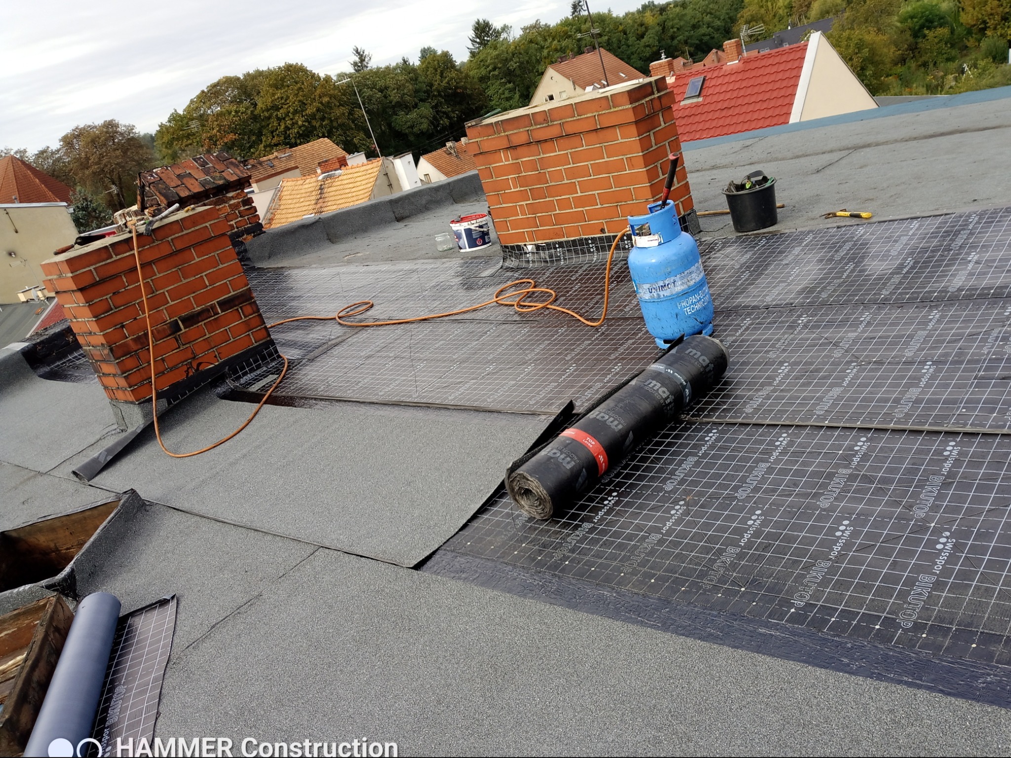 Widok z góry na dach w trakcie pokrywania papą termozgrzewalną z widocznymi dwoma kominami z cegły, butlą gazową, narzędziami i rolką papy. Na dachu widoczne są również inne budynki.
