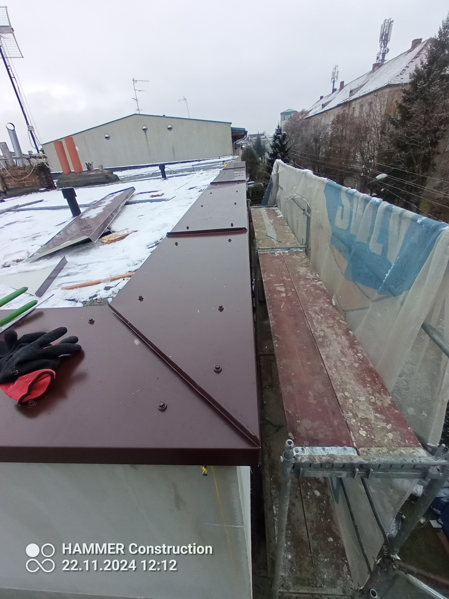 Fragment dachu pokrytego nową, brązową blachą trapezową, widoczne narzędzia dekarskie i rękawice robocze, w tle rusztowanie i zabudowa miejska w zimowej scenerii.