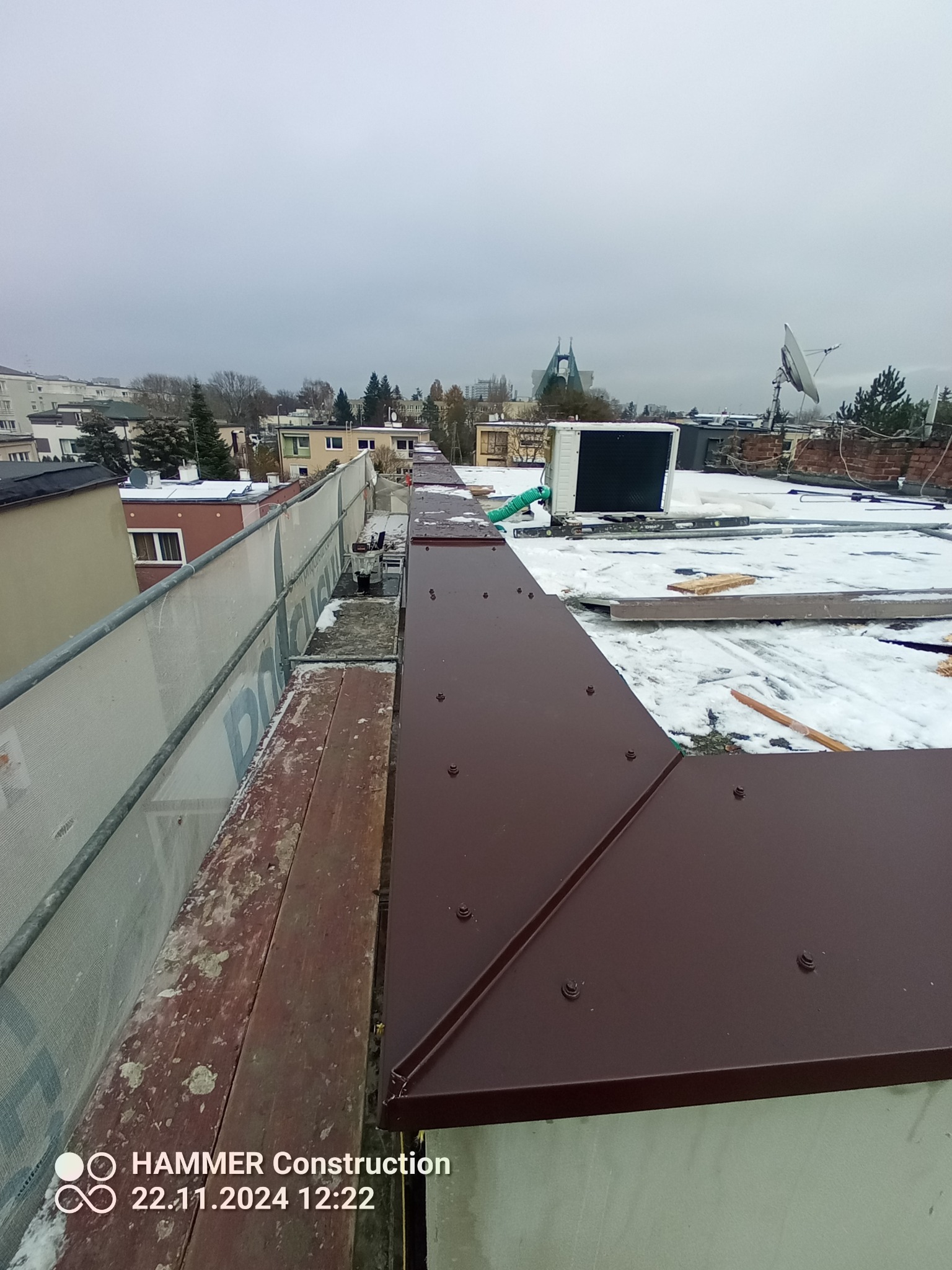 Montaż brązowych obróbek blacharskich na płaskim dachu budynku w zimowej scenerii, widok z rusztowania, elementy wentylacji i instalacji na dachu w tle.