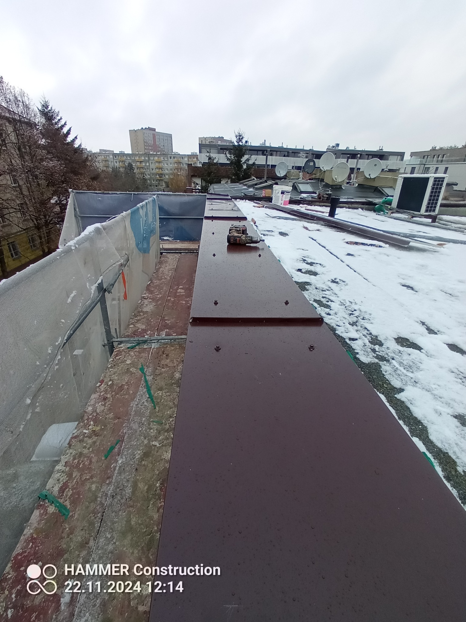 Widok z góry na fragment dachu z brązowymi elementami blacharskimi, częściowo pokrytego śniegiem, z widocznymi budynkami mieszkalnymi w tle i zabezpieczeniem rusztowania po lewej stronie.