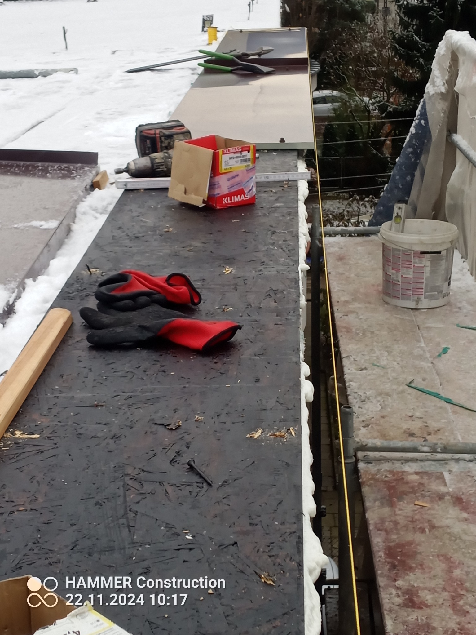 Prace dekarskie w zimowych warunkach: narzędzia, wkrętarka, puszka z wkrętami Klimas, rękawice robocze i poziomica na dachu pokrytym śniegiem, widoczne pianowanie krawędzi.