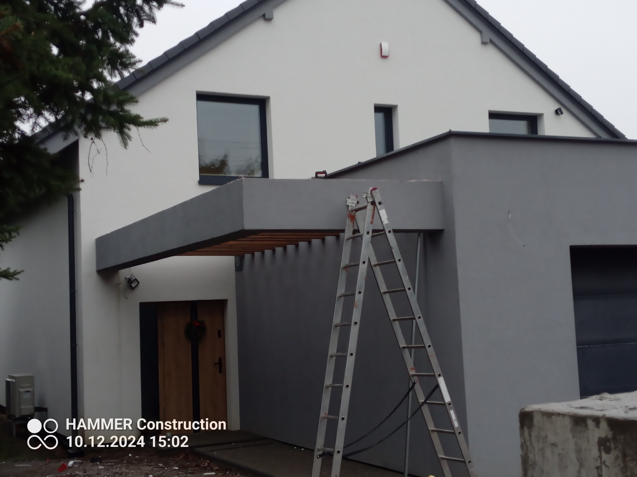 Nowoczesny dom z szarą elewacją i drewnianą pergolą, widoczna drabina oparta o ścianę, zima