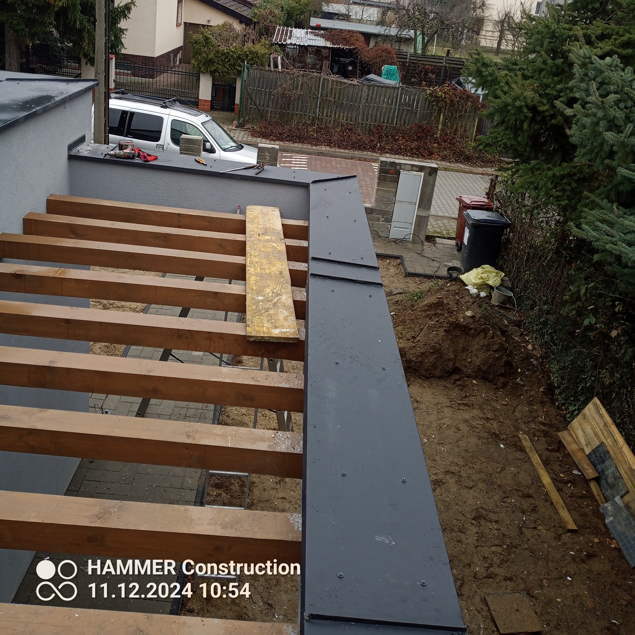 Konstrukcja dachu w trakcie budowy z widocznymi drewnianymi belkami i fragmentem obróbki blacharskiej w kolorze grafitowym, na tle zabudowań i zieleni.