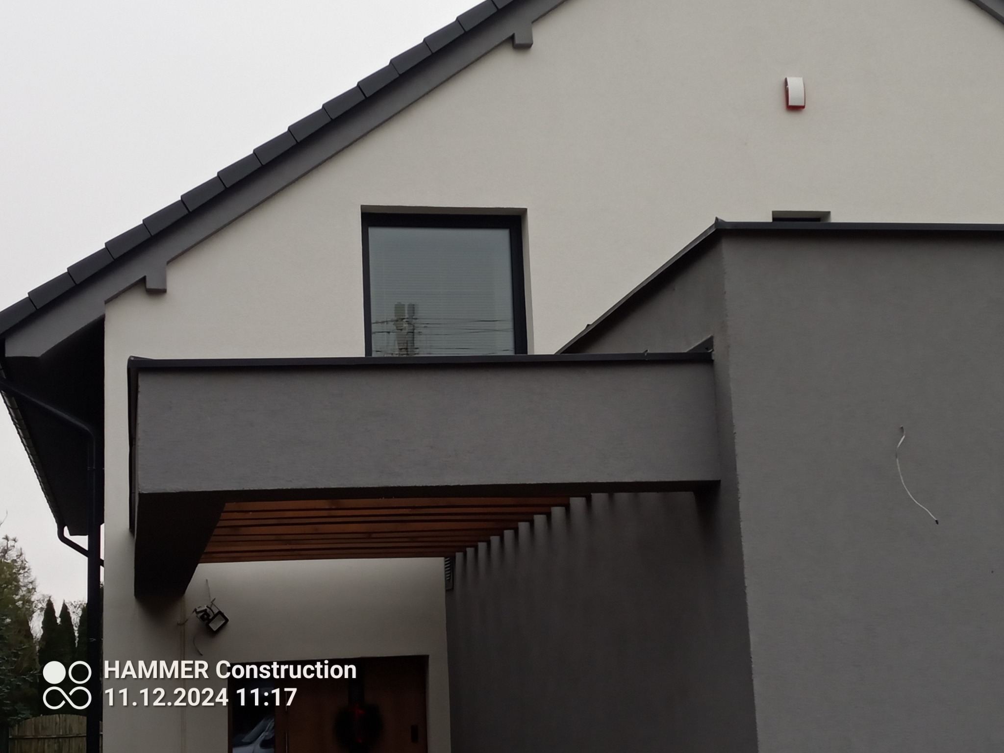 Dwukondygnacyjny dom z szarym dachem i drewnianą pergolą nad wejściem, widoczny fragment rynny i okno z żaluzjami.