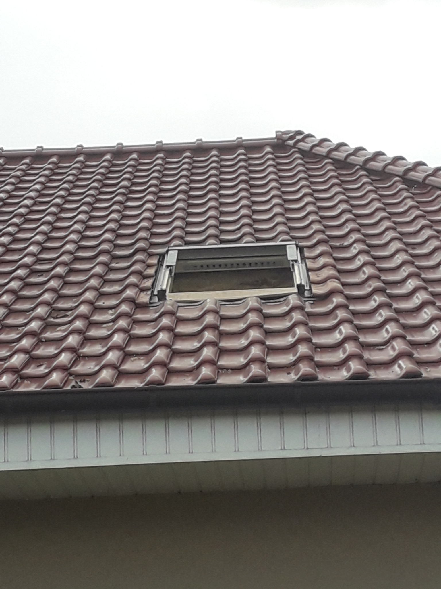 Czerwona dachówka ceramiczna z zamontowanym oknem dachowym, widoczne uszczelnienia i fragment elewacji budynku w tle.