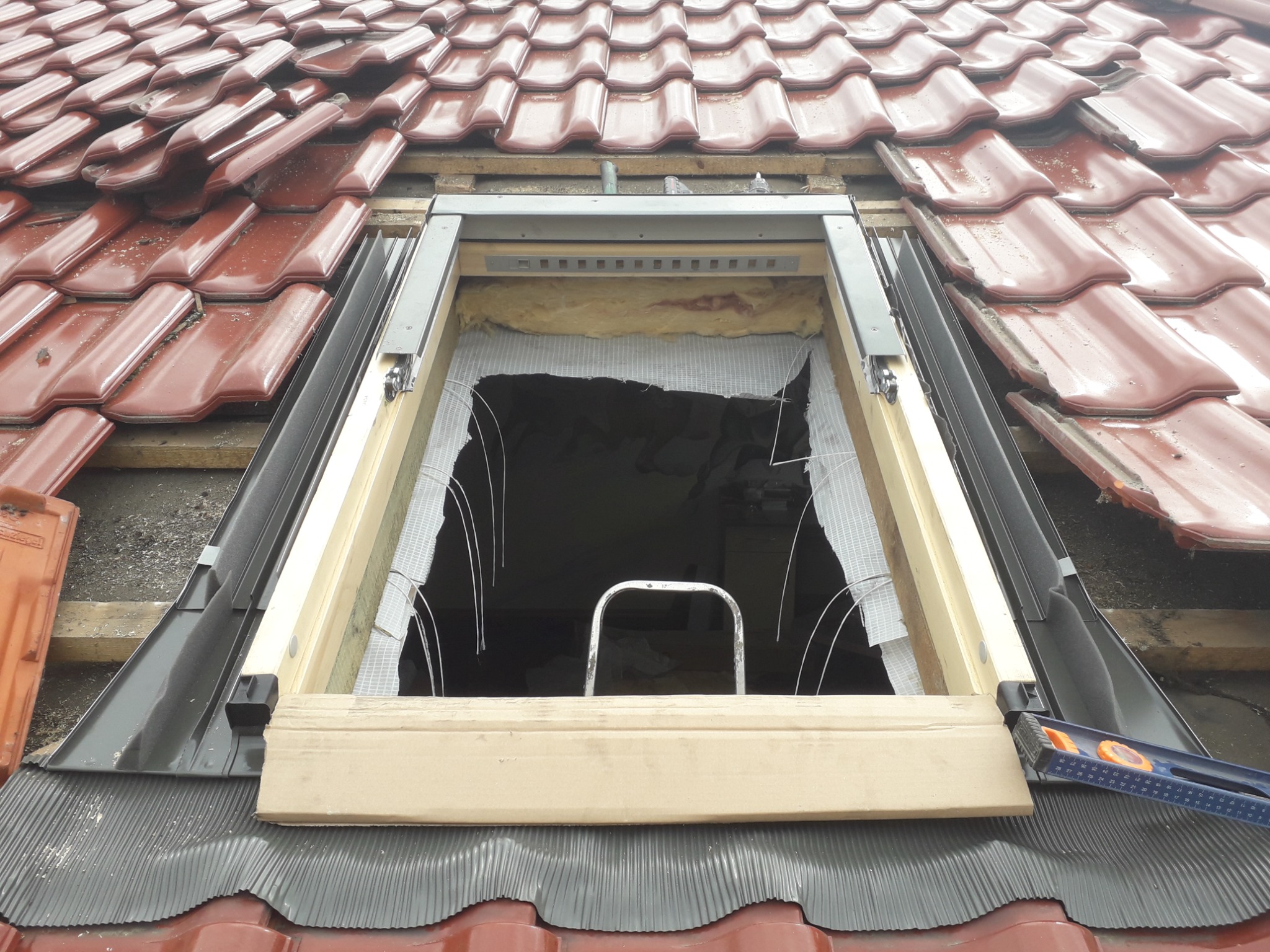 Montaż okna dachowego na czerwonej dachówce, widoczne elementy konstrukcyjne, izolacja i poziomica.
