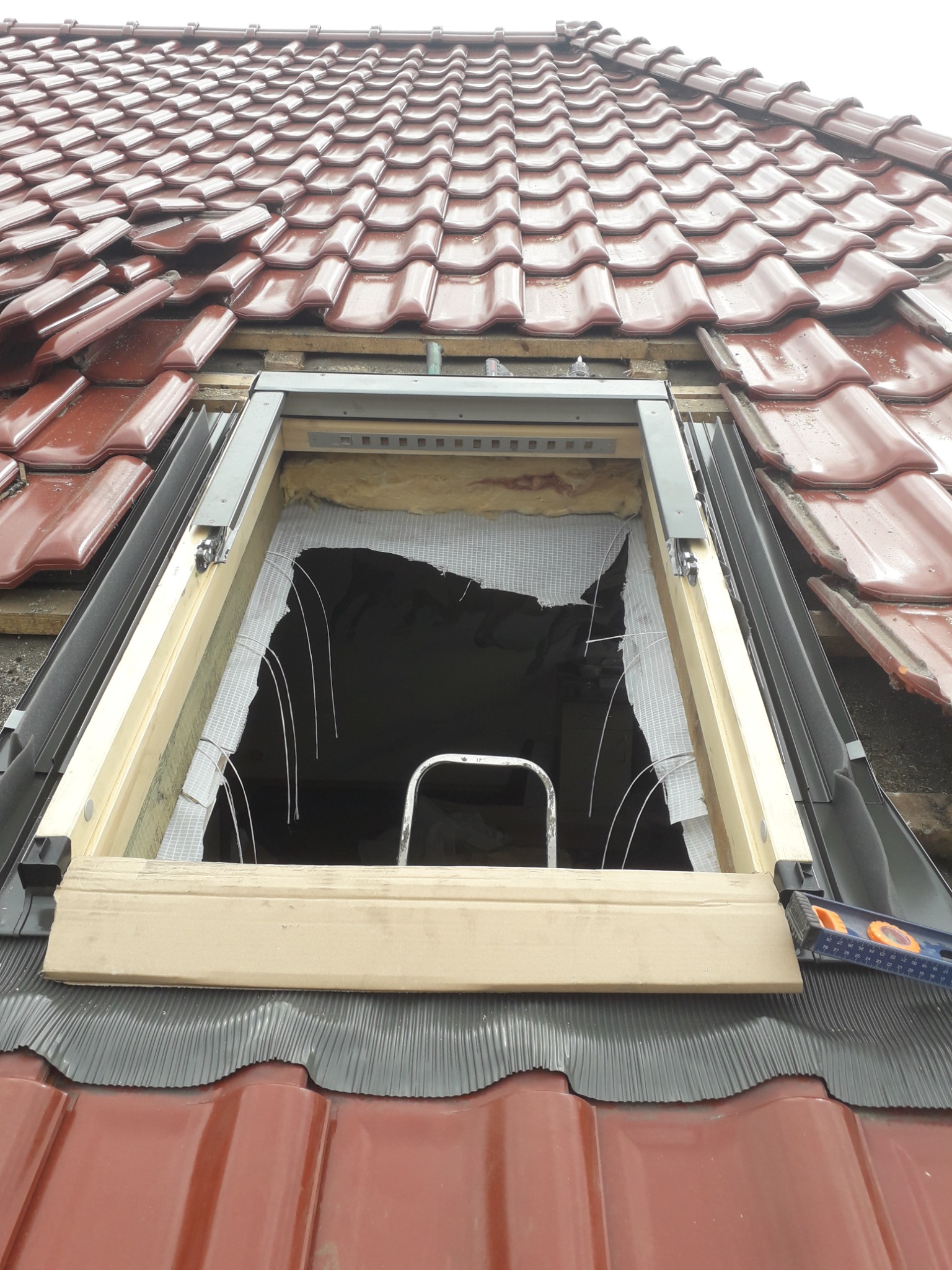 Montaż okna dachowego w czerwonej dachówce ceramicznej, widoczne elementy konstrukcyjne, izolacja i poziomica.