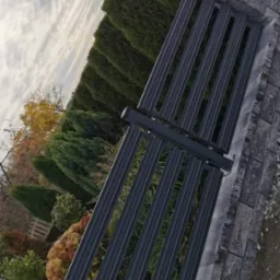 Nowoczesna, metalowa brama posesji w Krzepicach, z pionowymi profilami, osadzona na kamiennych słupkach i podmur&oacute;wce, na tle zieleni i pochmurnego nieba.