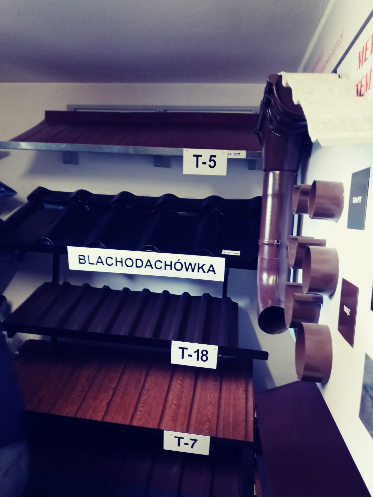 Ekspozycja różnych materiałów na dach: blachodachówka, blacha trapezowa T-7 i T-18, dachówka oraz system odprowadzania wody deszczowej z rynną i rurami spustowymi w kolorze brązowym.