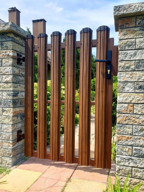 Brama ogrodowa z brązowych, pionowych paneli, osadzona między kamiennymi słupkami. Widoczny czarny uchwyt i fragment ścieżki z kostki brukowej.