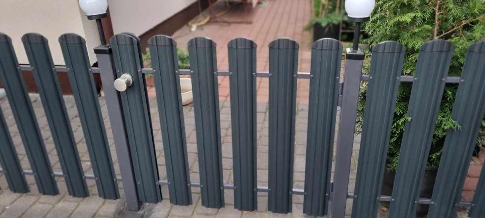 Nowoczesna, grafitowa furtka z pionowych paneli, z okrągłą gałką, oświetlona kulistą lampą, prowadząca do posesji z kostki brukowej i zielenią.