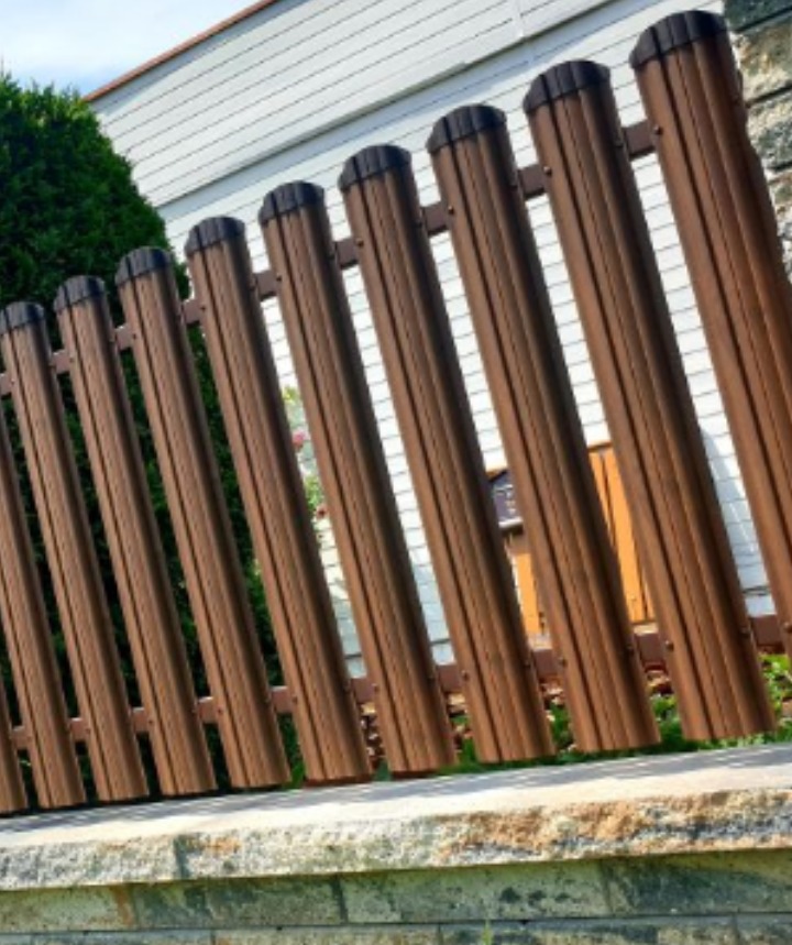 Nowoczesne, brązowe ogrodzenie panelowe z metalowych profili, zamontowane na kamiennej podmurówce, z widocznym fragmentem białej elewacji domu w tle.